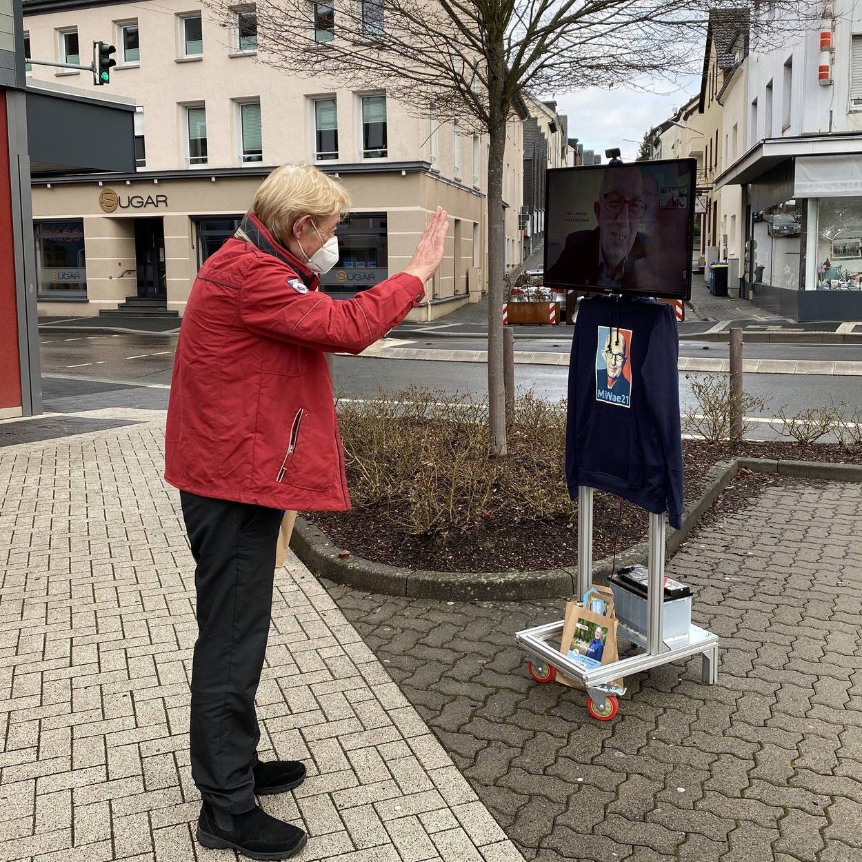 A wheeled screen with a battery and mobile data connection shows a member of parliament joining by video call, while a passer-by waves to it on a market square